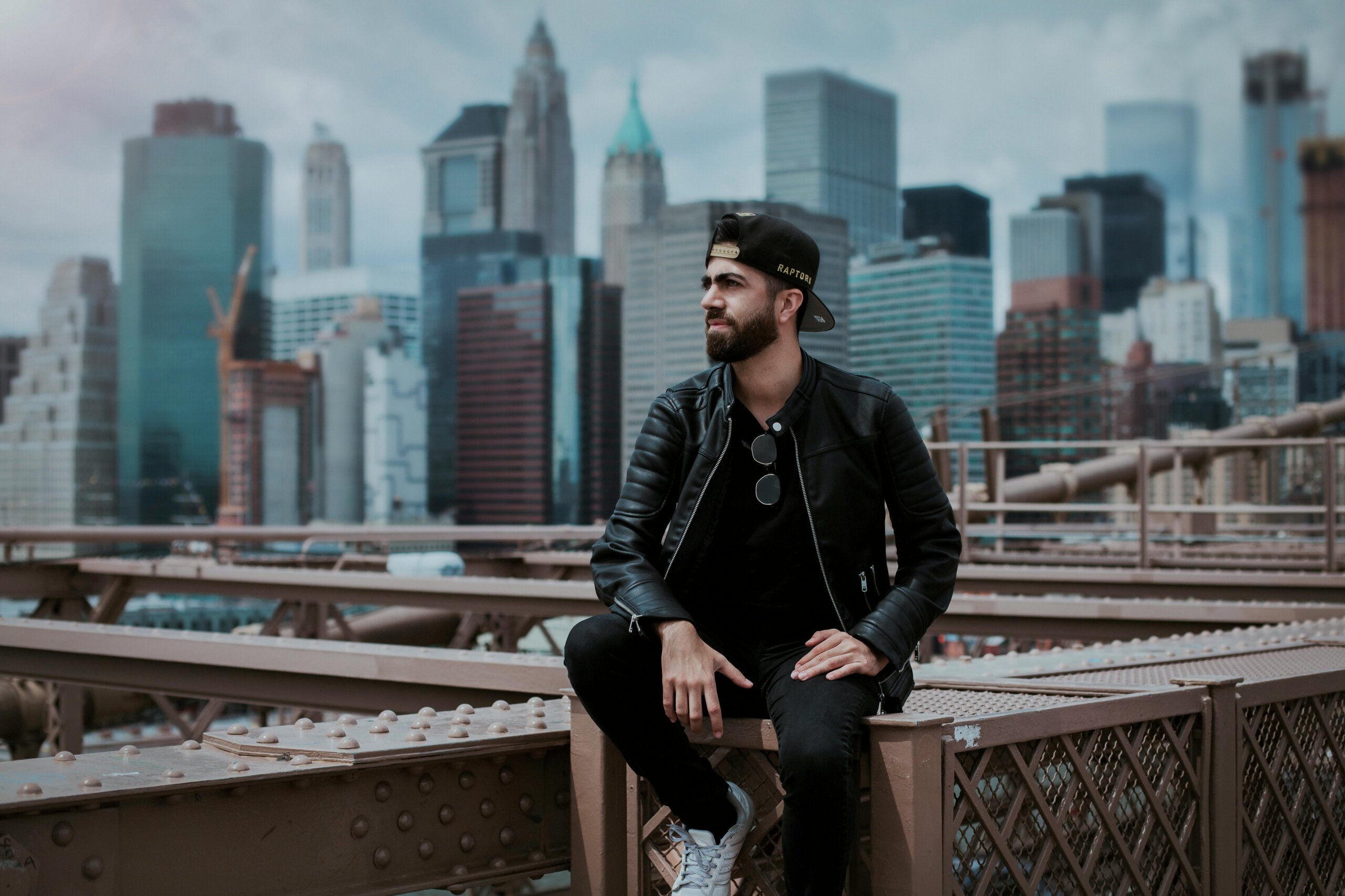 A man wearing a black leather jacket sitting on Brooklyn Bridge with New York City skyline in the background.
